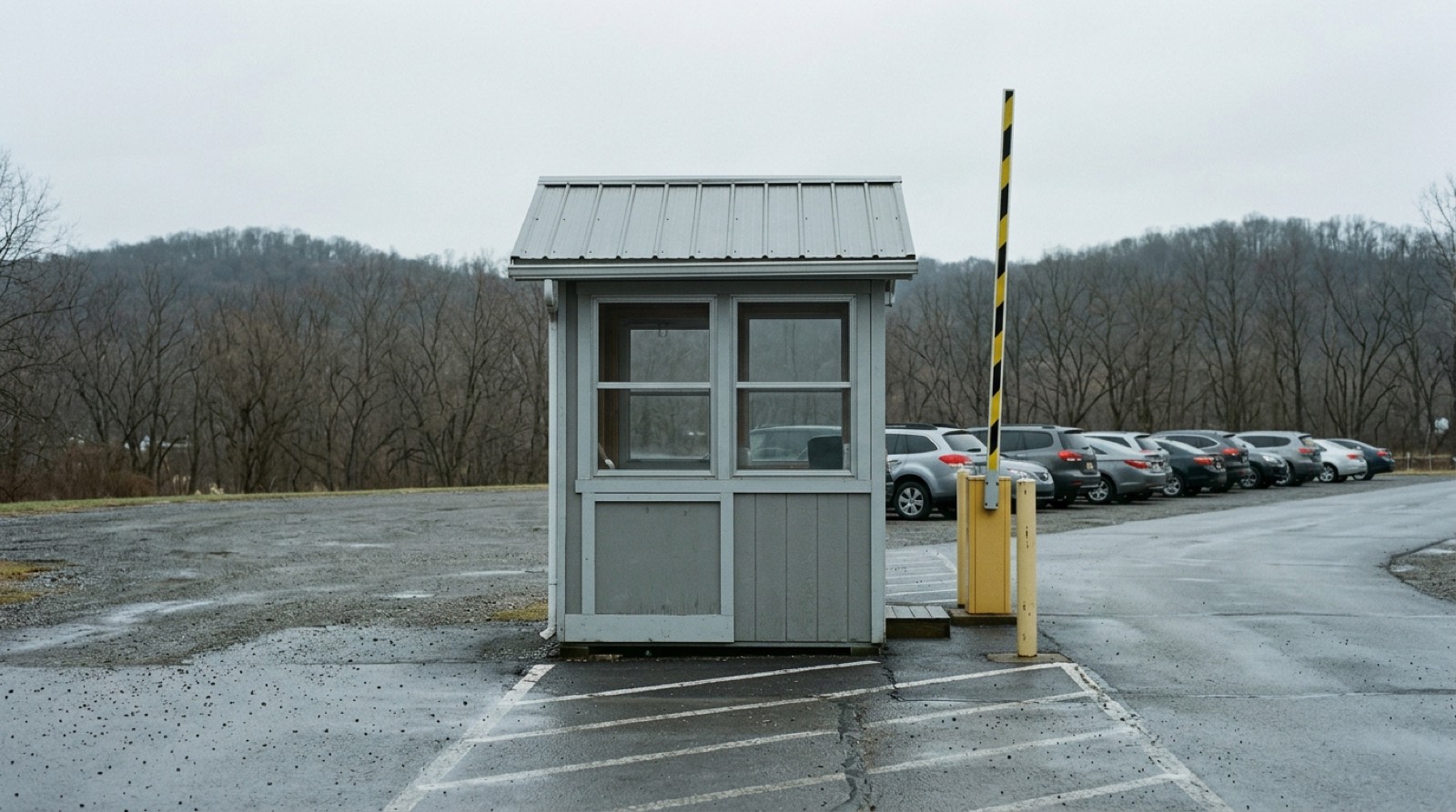 A small empty attendant booth at the head of a parking row, with a raised barrier arm and wet asphalt around it, in flat gray light