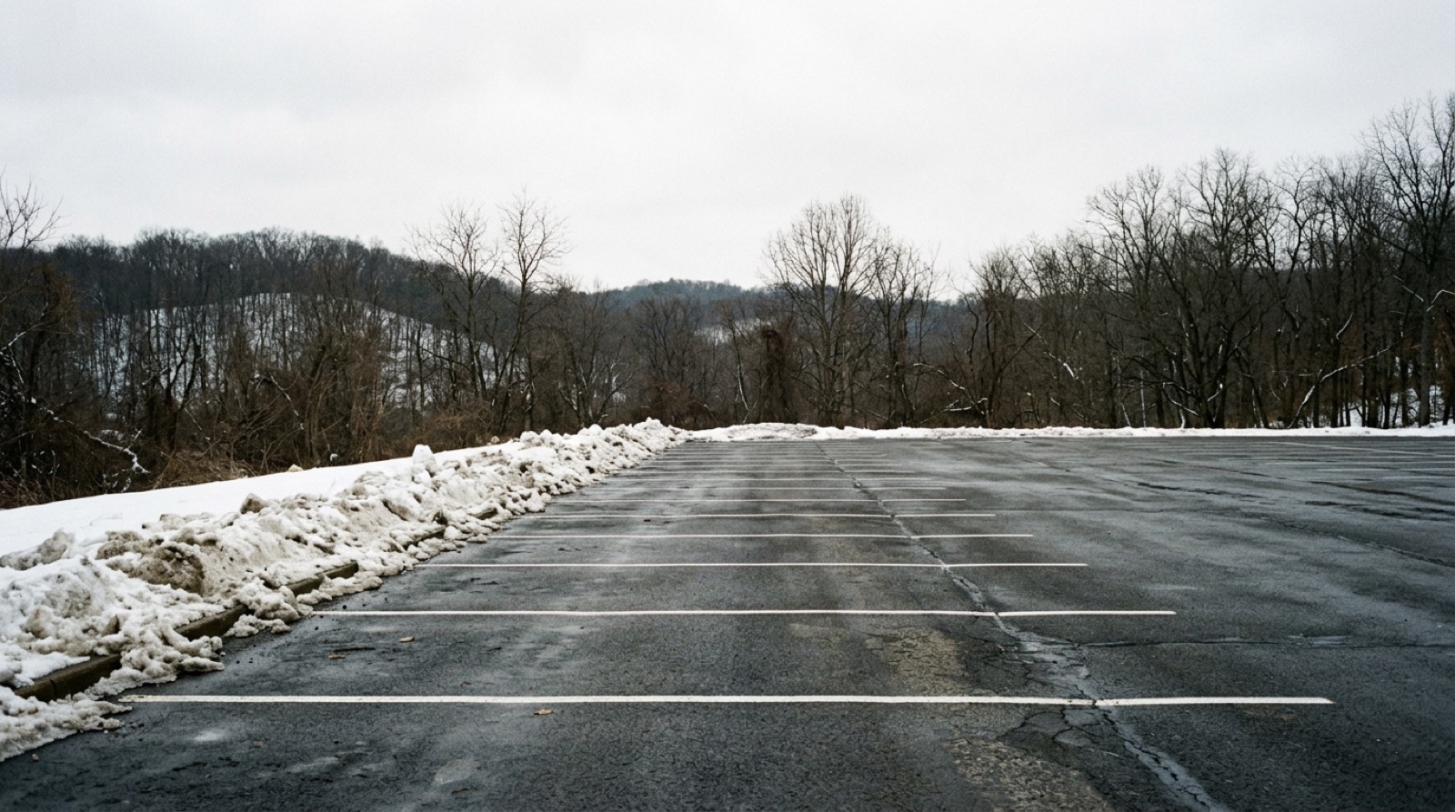 A plowed parking row with banked snow along one side and clear painted lines on the asphalt, photographed square on under a white winter sky