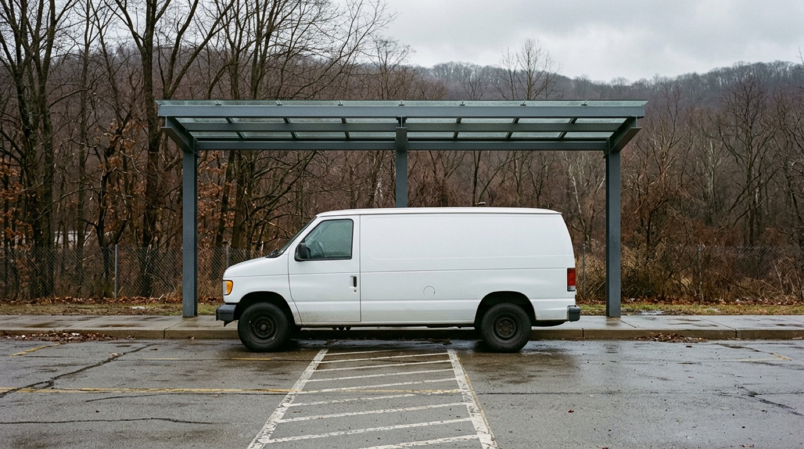 A plain white shuttle van standing empty at a marked curb under a simple steel canopy, seen square on in flat daylight, with no lettering on it